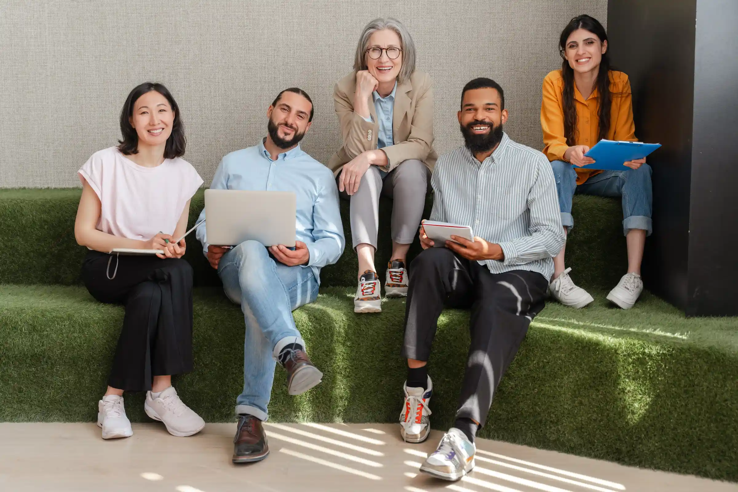 The image shows a group of five people sitting together on tiered green seating in what appears to be a modern, relaxed office or collaborative workspace. They are all smiling and seem comfortable, giving the impression of a friendly and diverse team.
